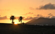 Die Sonne geht am Horizont zwischen 2 Palmen unter und im Abendrot sieht man ländliche Häuser vor Bergen stehen