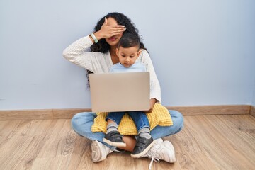 Poster - Young hispanic mother and kid using computer laptop sitting on the floor smiling and laughing with hand on face covering eyes for surprise. blind concept.