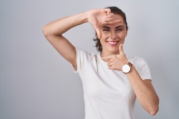 Poster - Beautiful brunette woman standing over isolated background smiling making frame with hands and fingers with happy face. creativity and photography concept.