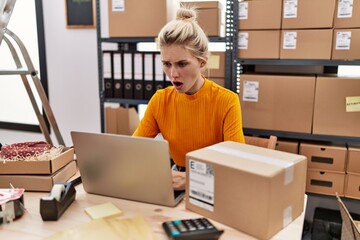 Canvas Print - Young blonde woman working at small business ecommerce using laptop scared and amazed with open mouth for surprise, disbelief face