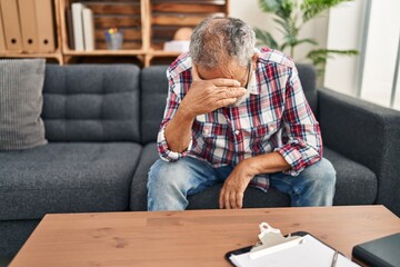 Wall Mural - Senior grey-haired man patient stressed with hands on face sitting on sofa at psychology clinic