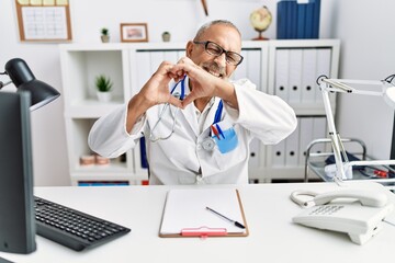 Wall Mural - Mature doctor man at the clinic smiling in love doing heart symbol shape with hands. romantic concept.