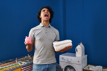 Wall Mural - Hispanic man with curly hair holding clean towels and piggy bank angry and mad screaming frustrated and furious, shouting with anger looking up.