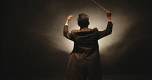 Unrecognizable Male Orchestra Conductor Controlling Music In Orchestra Pit By Movement Of His Hands And White Baton, Studio Shot On Black Background 