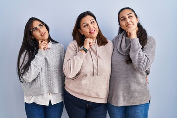 Wall Mural - Mother and two daughters standing over blue background with hand on chin thinking about question, pensive expression. smiling with thoughtful face. doubt concept.