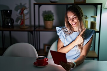 Canvas Print - Young hispanic woman using touchpad sitting on the table at night cheerful with a smile on face pointing with hand and finger up to the side with happy and natural expression