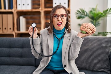 Sticker - Middle age hispanic woman working at therapy office holding brain clueless and confused expression. doubt concept.