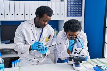 Canvas Print - African american man and woman scientists using microscope writing on document at laboratory