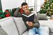 © Krakenimages.com - Young hispanic man reading book sitting on sofa by christmas tree at home