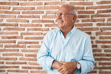 Poster - Senior man with grey hair standing over bricks wall looking away to side with smile on face, natural expression. laughing confident.