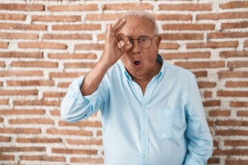 Poster - Senior man with grey hair standing over bricks wall doing ok gesture shocked with surprised face, eye looking through fingers. unbelieving expression.