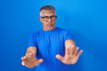 Poster - Hispanic man with grey hair standing over blue background moving away hands palms showing refusal and denial with afraid and disgusting expression. stop and forbidden.