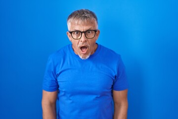 Canvas Print - Hispanic man with grey hair standing over blue background afraid and shocked with surprise and amazed expression, fear and excited face.