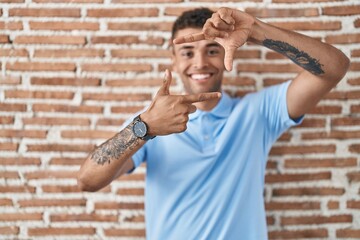 Sticker - Brazilian young man standing over brick wall smiling making frame with hands and fingers with happy face. creativity and photography concept.