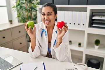 Canvas Print - Young hispanic woman working at dietitian clinic holding green apple sticking tongue out happy with funny expression.