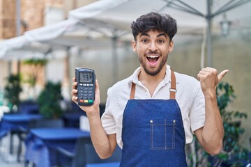 Poster - Arab man with beard wearing waiter apron at restaurant terrace holding dataphone pointing thumb up to the side smiling happy with open mouth