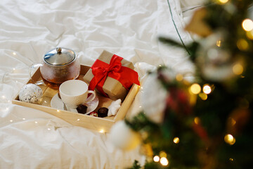 Wall Mural - Christmas concept - wooden tray with cup, teapot, candies and gift box on the bed near decorated Christmas tree