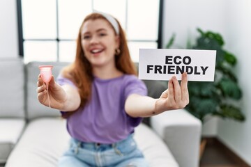 Wall Mural - Young redhead woman smiling confident holding menstrual cup and eco friendly banner at home