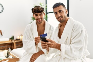 Wall Mural - Two hispanic men couple holding credit card and smartphone sitting on massage table at beauty center