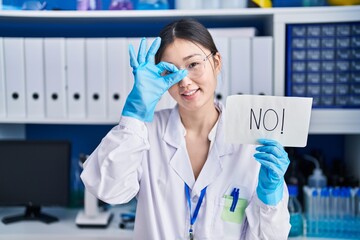 Wall Mural - Chinese young woman working at scientist laboratory holding no banner smiling happy doing ok sign with hand on eye looking through fingers