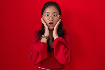 Wall Mural - Asian young woman standing over red background afraid and shocked, surprise and amazed expression with hands on face