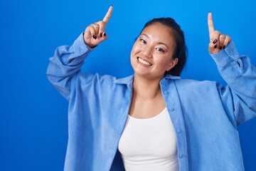 Wall Mural - Asian young woman standing over blue background smiling amazed and surprised and pointing up with fingers and raised arms.