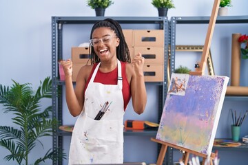 Canvas Print - Young african american with braids at art studio painting on canvas very happy and excited doing winner gesture with arms raised, smiling and screaming for success. celebration concept.