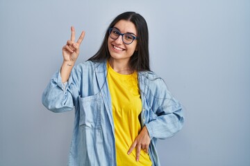 Canvas Print - Young hispanic woman standing over blue background smiling looking to the camera showing fingers doing victory sign. number two.