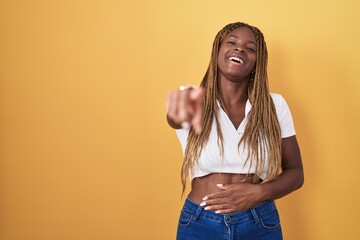 Wall Mural - African american woman with braided hair standing over yellow background laughing at you, pointing finger to the camera with hand over body, shame expression