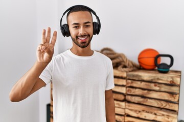 Poster - African american man listening to music using headphones at the gym showing and pointing up with fingers number three while smiling confident and happy.