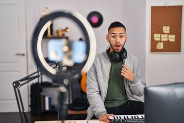 Poster - Young hispanic man playing piano at music studio recording himself scared and amazed with open mouth for surprise, disbelief face