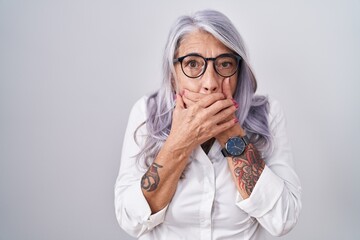 Wall Mural - Middle age woman with tattoos wearing glasses standing over white background shocked covering mouth with hands for mistake. secret concept.