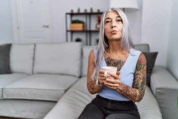 Wall Mural - Middle age grey-haired woman drinking coffee sitting on the sofa at home looking at the camera blowing a kiss on air being lovely and sexy. love expression.