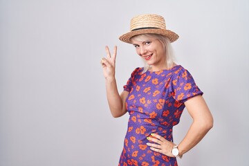 Poster - Young caucasian woman wearing flowers dress and summer hat smiling looking to the camera showing fingers doing victory sign. number two.