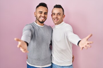 Poster - Homosexual couple standing over pink background looking at the camera smiling with open arms for hug. cheerful expression embracing happiness.