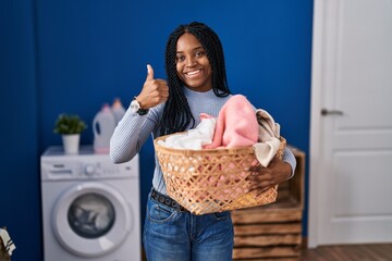 Wall Mural - African american woman holding laundry basket smiling happy and positive, thumb up doing excellent and approval sign