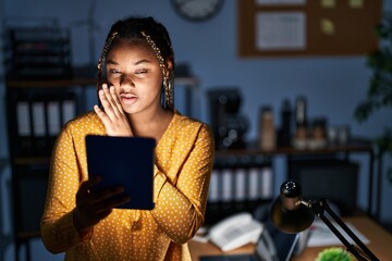 Wall Mural - African american woman with braids working at the office at night with tablet hand on mouth telling secret rumor, whispering malicious talk conversation