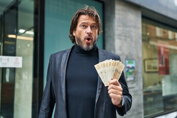 Canvas Print - Handsome middle age man holding 100 danish krone banknotes scared and amazed with open mouth for surprise, disbelief face