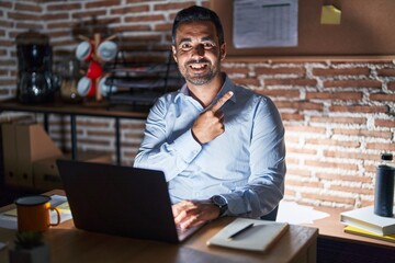 Canvas Print - Hispanic man with beard working at the office at night cheerful with a smile on face pointing with hand and finger up to the side with happy and natural expression