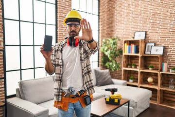 Canvas Print - Young hispanic man with beard working at home renovation holding smartphone with open hand doing stop sign with serious and confident expression, defense gesture