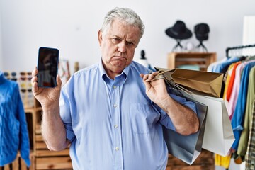 Wall Mural - Senior man with grey hair holding shopping bags and showing smartphone screen depressed and worry for distress, crying angry and afraid. sad expression.