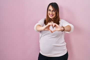 Poster - Pregnant woman standing over pink background smiling in love doing heart symbol shape with hands. romantic concept.