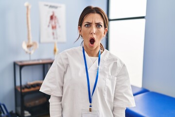 Canvas Print - Young brunette woman working at pain recovery clinic in shock face, looking skeptical and sarcastic, surprised with open mouth