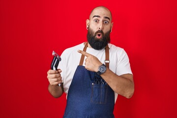 Canvas Print - Young hispanic man with beard and tattoos wearing barber apron holding razor surprised pointing with finger to the side, open mouth amazed expression.