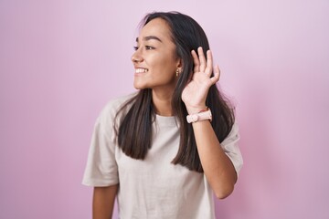 Wall Mural - Young hispanic woman standing over pink background smiling with hand over ear listening an hearing to rumor or gossip. deafness concept.