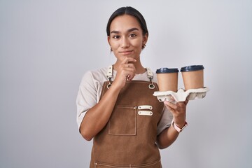 Wall Mural - Young hispanic woman wearing professional waitress apron holding coffee looking confident at the camera smiling with crossed arms and hand raised on chin. thinking positive.