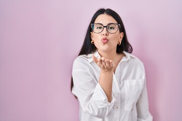 Sticker - Young brunette woman standing over pink background looking at the camera blowing a kiss with hand on air being lovely and sexy. love expression.