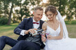 Smiling and shy groom and bride drinking and pouring bubbly champagne wine from bottle to glasses together, sit in park
