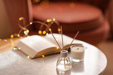 Wall Mural - Cozy home atmosphere with liquid fragrance in glass bottle and sticks with open paper book over glow lights on marble table in living room close up. Winter holiday season.