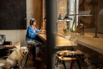 Wall Mural - Woman sits with her cute adorable dog at modern coffee shop and works on laptop. Pet friendly places and modern digital lifestyle concept